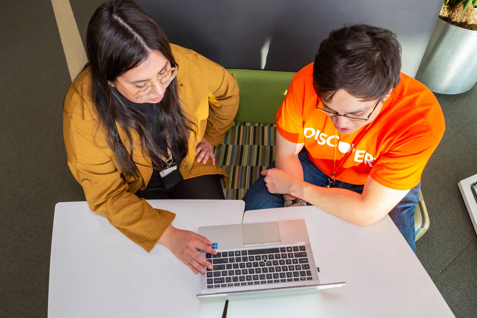 An overview perspective of two people sitting at a white table looking at a laptop. A person&#x27;s shirt reads, “DISCOVER.”