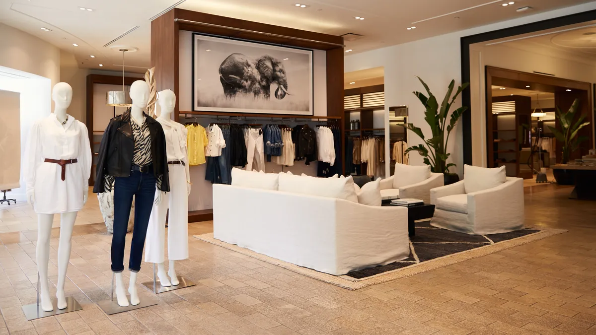 The interior of a store features mannequins to the left, a rack of clothes in the middle, a large plant to the right, and furniture in the middle.