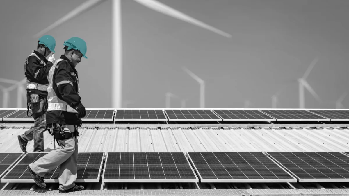 Two workers on a rooftop covered with solar panels, with wind turbines in the background.