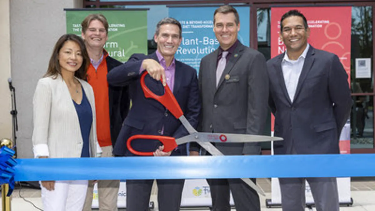 A group of people pose with a giant pair of red-handled scissors cutting a blue ribbon in front of Firmenich's new building