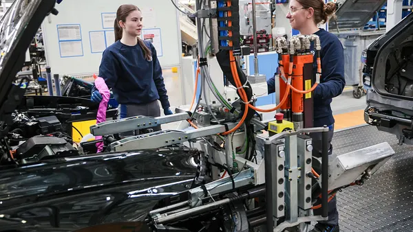 Two women work complex machinery to aid in the building of a car.