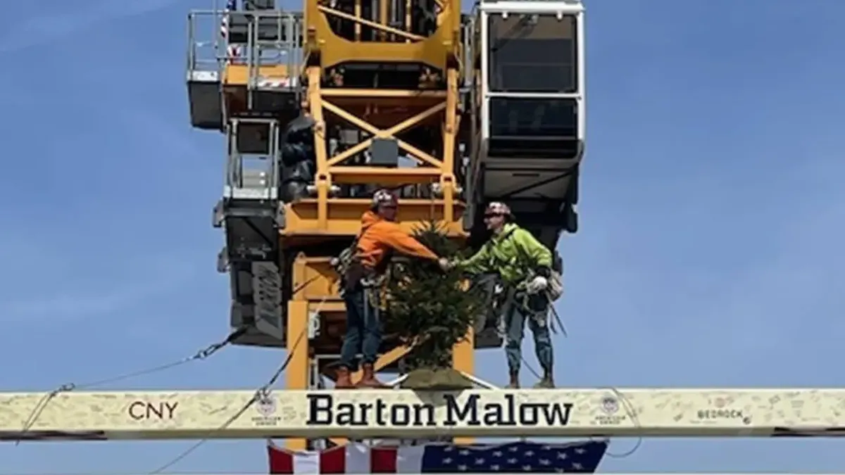 Workers in front of heavy equipment stand with a beam that reads "Barton Malow" on top of a tall building.