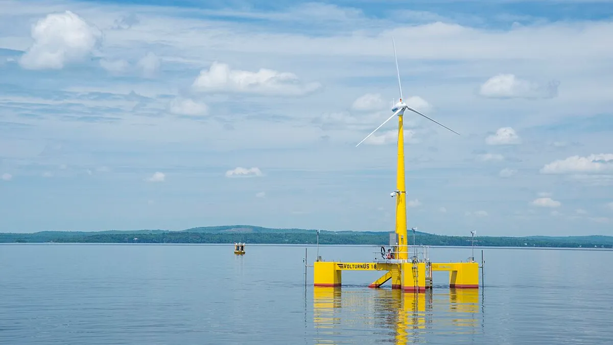 A floating wind turbine in still ocean water with wooded coast in the background.