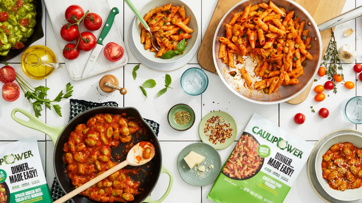 Three pans, each one containing a different variety of Caulipower's Frozen Meals, sit on a white tiled counter. Packages of the frozen meals and ingredients including tomatoes and herbs are nearby.