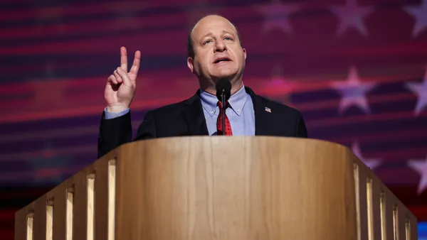 Jared Polis stands behind a podium on a stage with an American flag projected behind him and holds up two fingers.