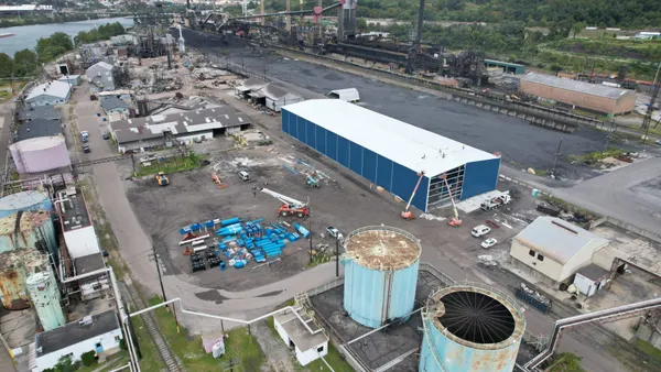 Aerial view of an industrial port with coal piles, several storage tanks and buildings, rail lines and a large industrial shed under construction. The complex sits on the bank of a river.