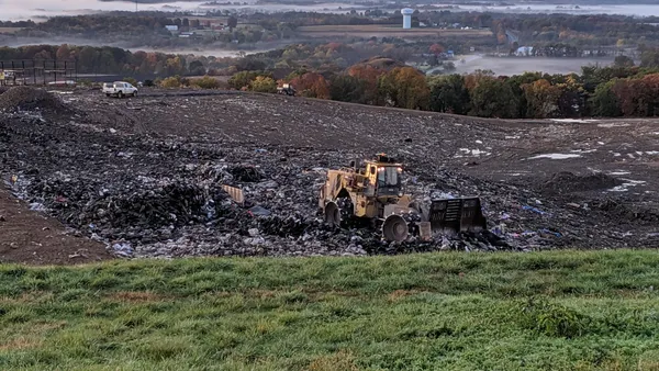 A bulldozer pushes trash on the open face of a landfill early in the morning. A town partially covered by fog is visible in the background.