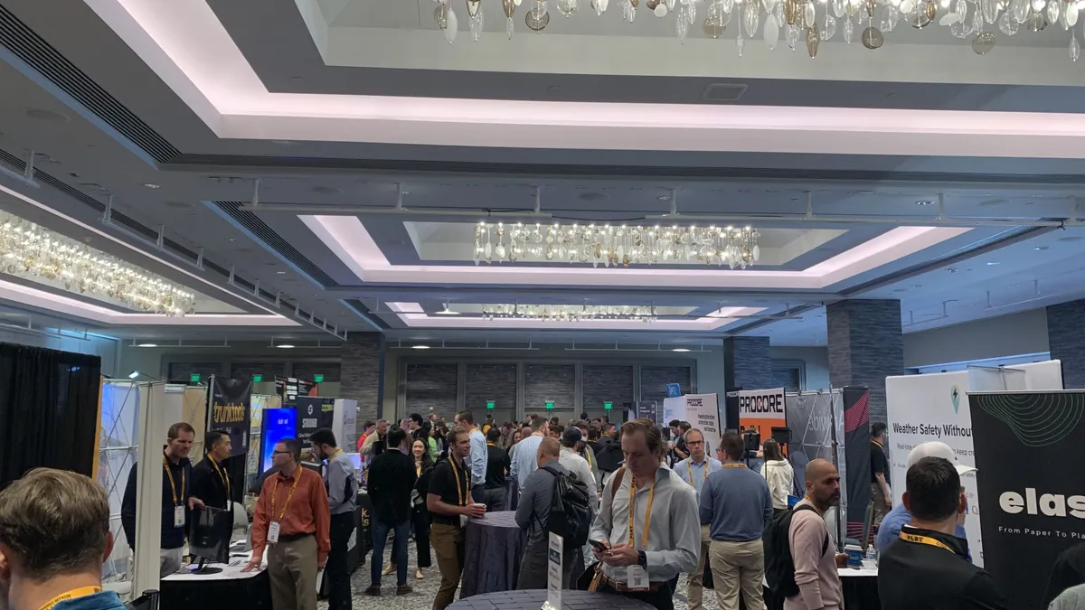 A crowd of people in business and business-casual clothes wander around an open space filled with booths for companies that represent construction technology firms.