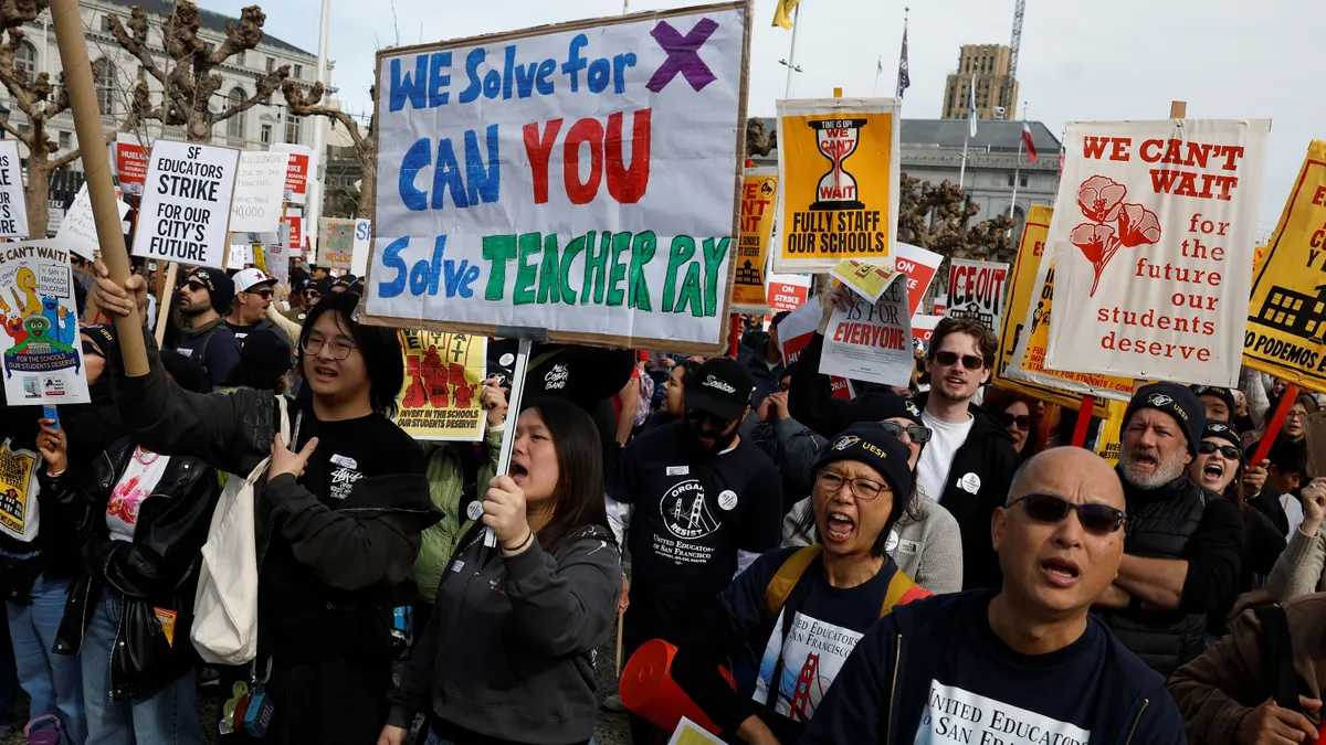 A large group of protesters stand together holding signs. One large sign in the center of the crowd says "We solve for X. Can you solve teacher pay?"