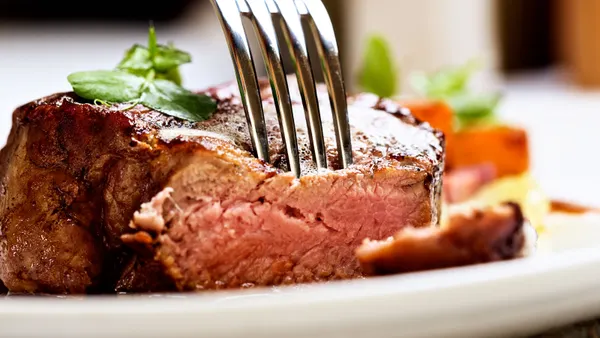 An image of a fork piercing a steak that sits on a white plate.