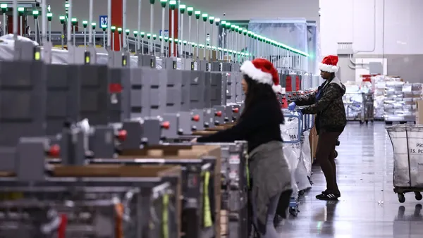 U.S. Postal Service employees in Santa hats sort packages inside a building.