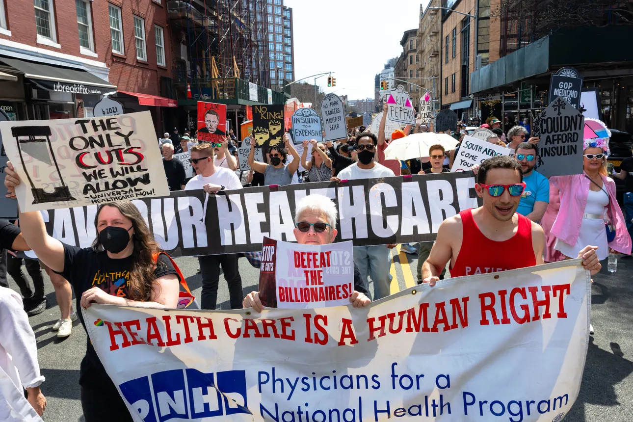 Three people hold a banner that says "Health Care is a Human Right" while others holding signs march behind them.