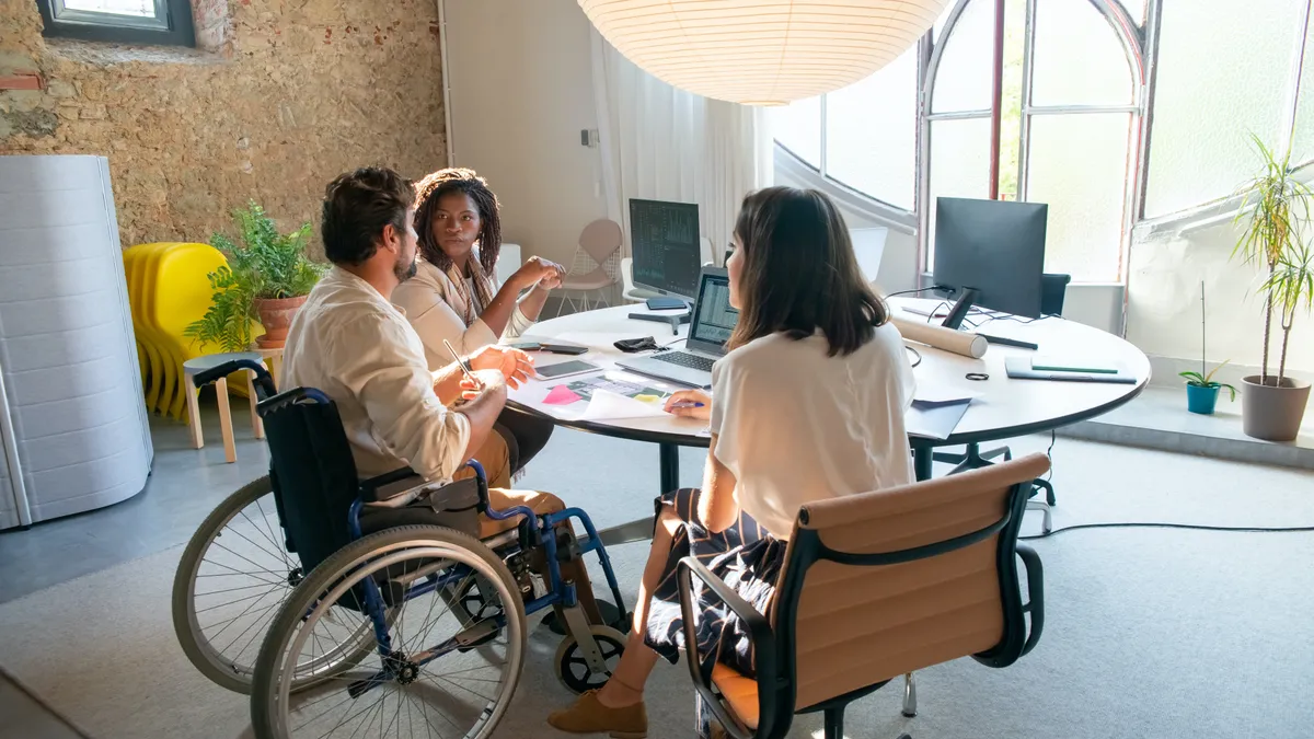 A group of coworkers sit at a table, discussing work. One of the coworkers is seated in his wheelchair.