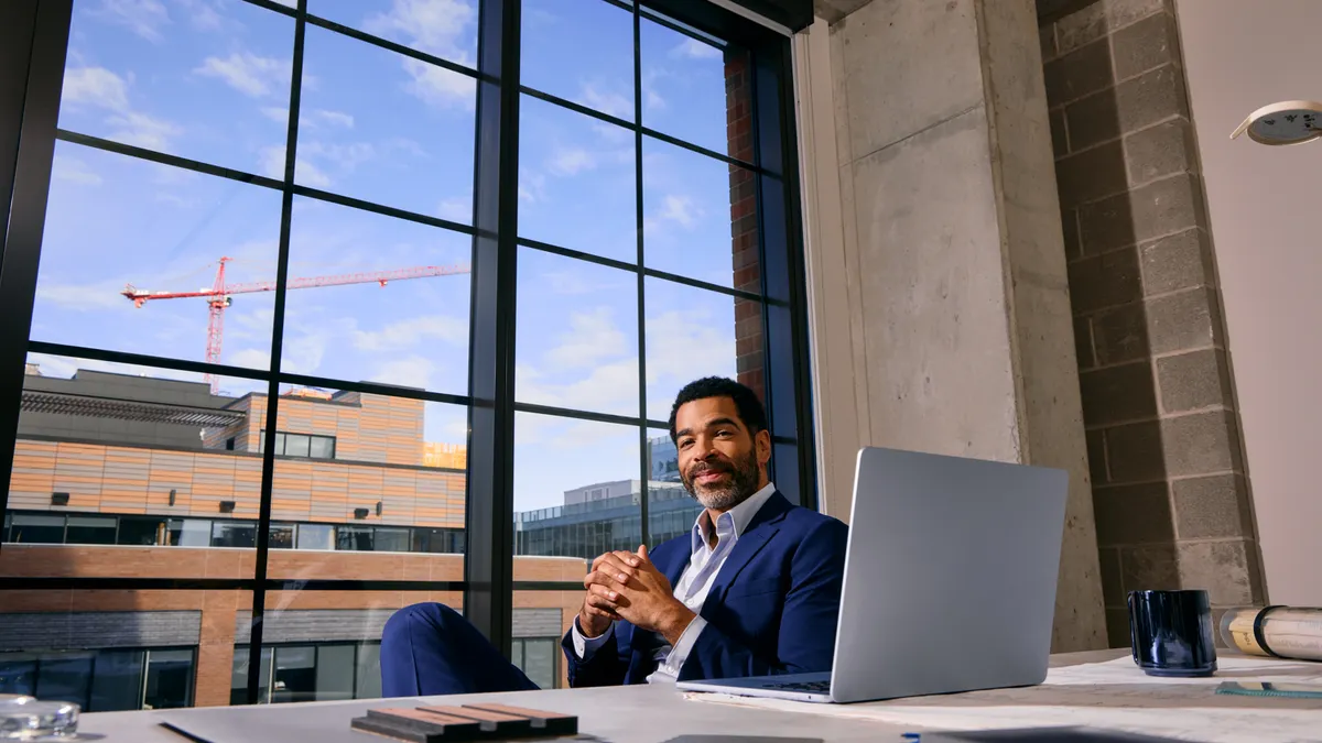 A confident man in a suit sits at a modern office desk near large windows, with buildings and a crane visible outside. The space feels professional and bright.