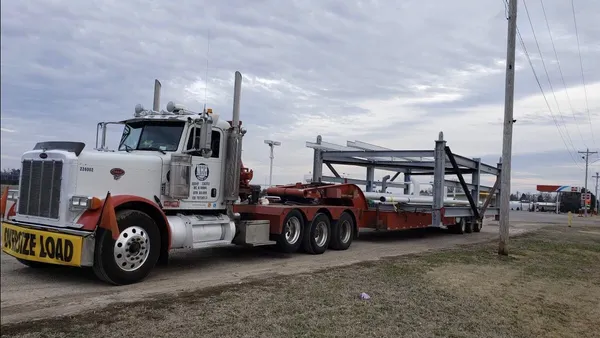 A truck transports a beam structure while displaying a yellow oversize load sign on the front of its cab.