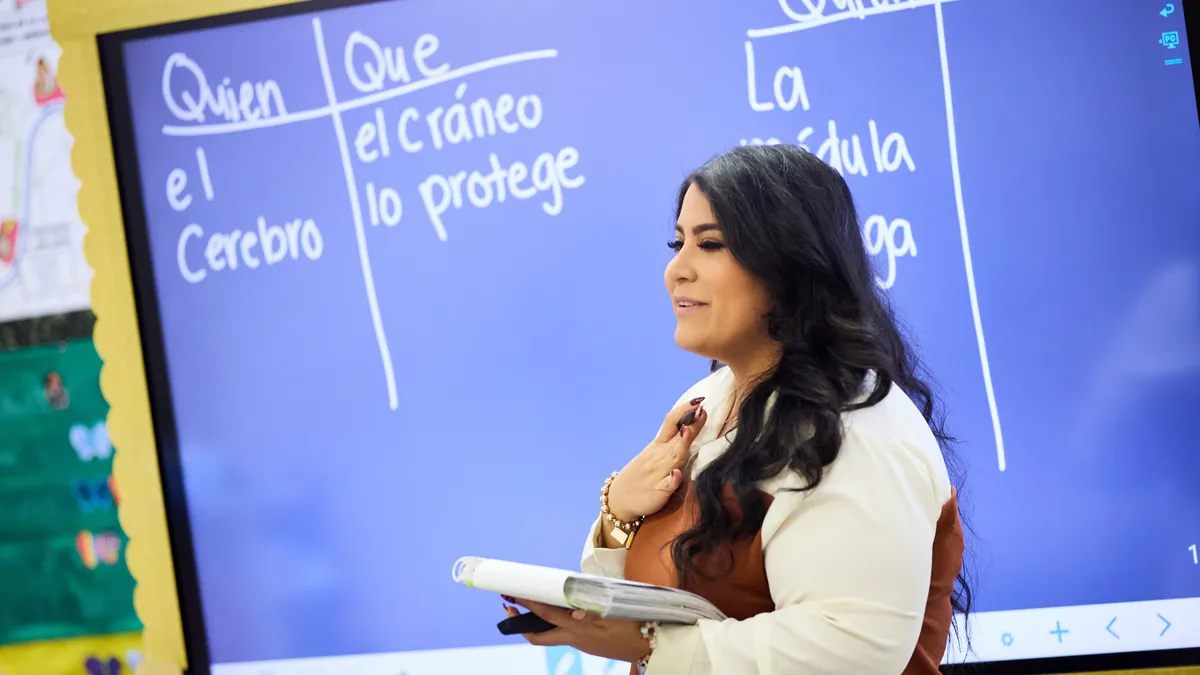 A woman teacher stands in front of a classroom with a smart board behind her.