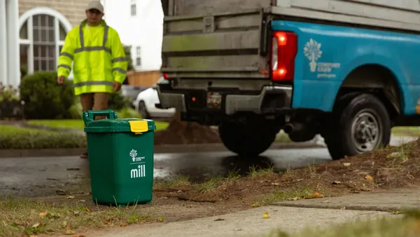 A small locking waste bin with the Compost Crew and Mill logos sits on a curb. A worker walks behind a collection truck toward the bin.