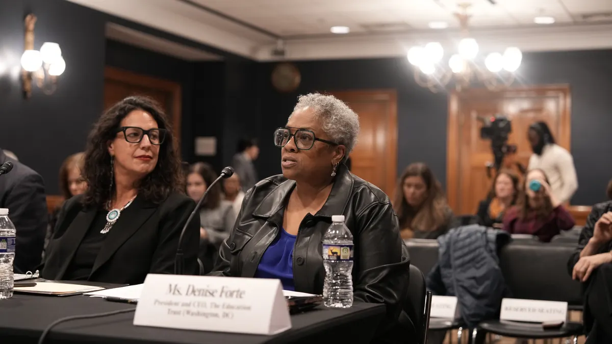 Two people are shown seated next to each other at a table in a large room. One person is talking and other people are seated in rows behind them.