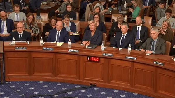 Six people in business clothes sit behind a dias in a congressional room.