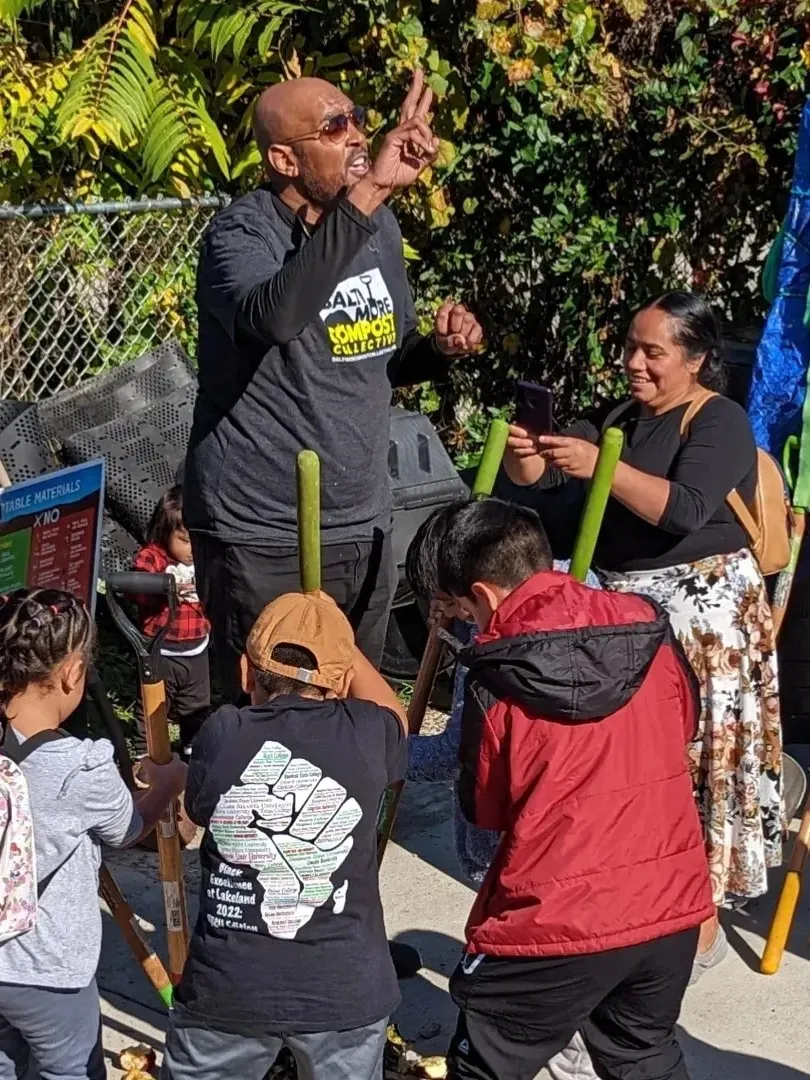 A man wearing a t-shirt with the Baltimore Compost Collective logo stands over children holding shovels.