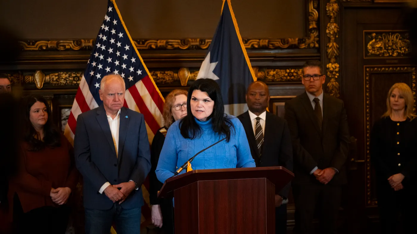 A person in a blue shirt speaks at a podium with people in suits and ties lined up behind her