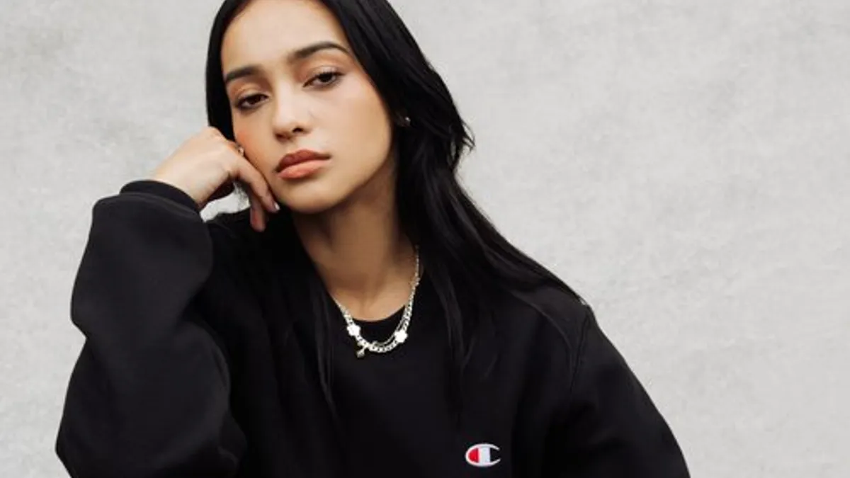 A model is seated on a green leather chair, wearing a black sweatshirt with the Champion logo.