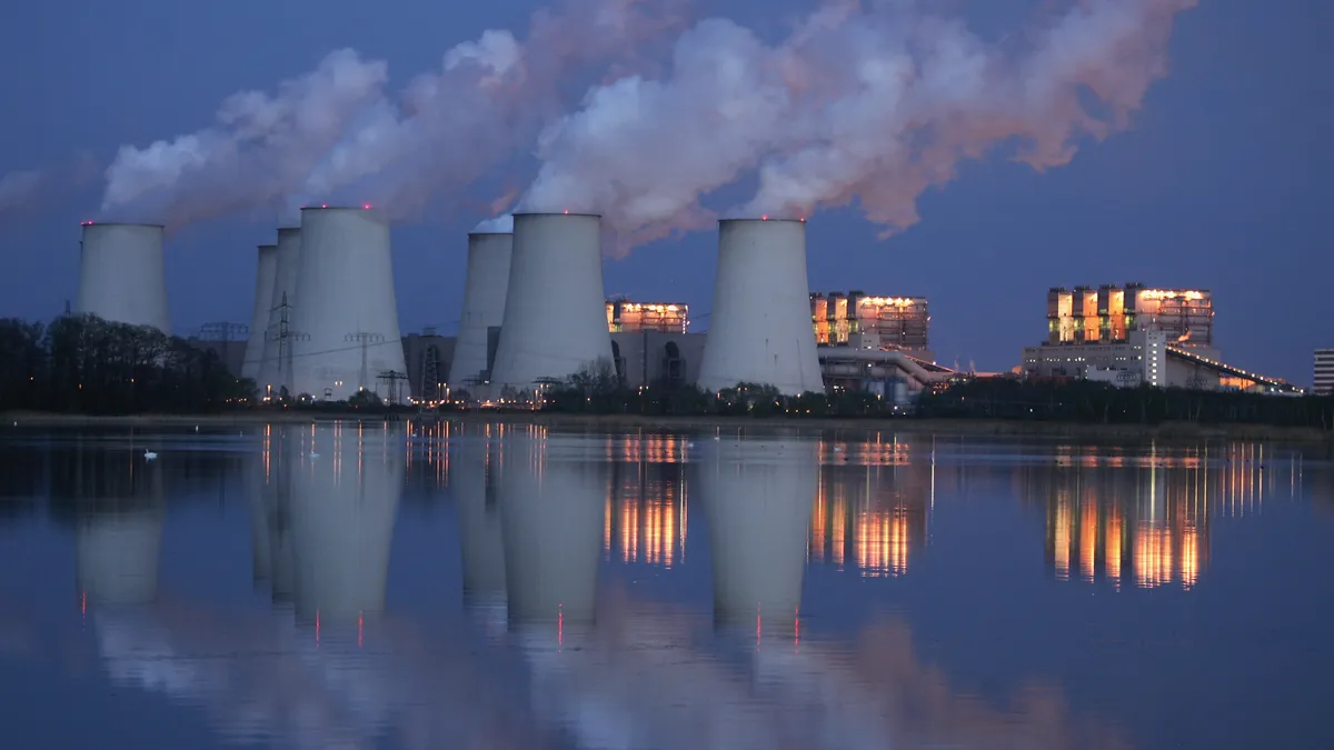 In the distance, exhaust plumes billow over seven power plants. The reflection of the power plants can be seen in a lake.