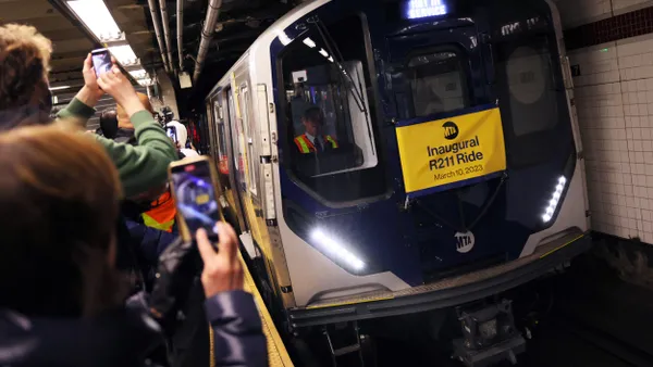 People on a subway platform hold up cell phones to photograph a new subway train arriving in the station with a sign announcing its inaugural ride