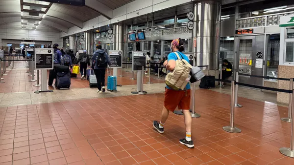 People with backpacks and luggage walk along a red tile floor toward buses seen though glass doors and windows.