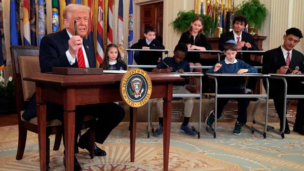 President Donald Trump sits at a desk and holds up a pen. Students are siting at desks behind the adult