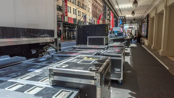 Production team places several professional boxes for musical equipment and costumes at the entrance of a Broadway theater with city buildings and a parking garage sign in the background.