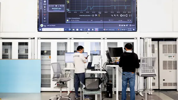 Two employees work at standing desks, facing a large screen.