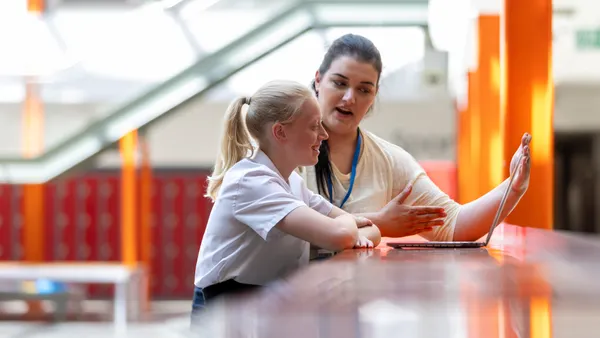 An adult is sitting at a table inside with a student. They are both looking at an open laptop.