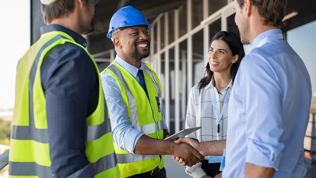 Four people, two in hard hats and vests, shake hands at a construction jobsite.