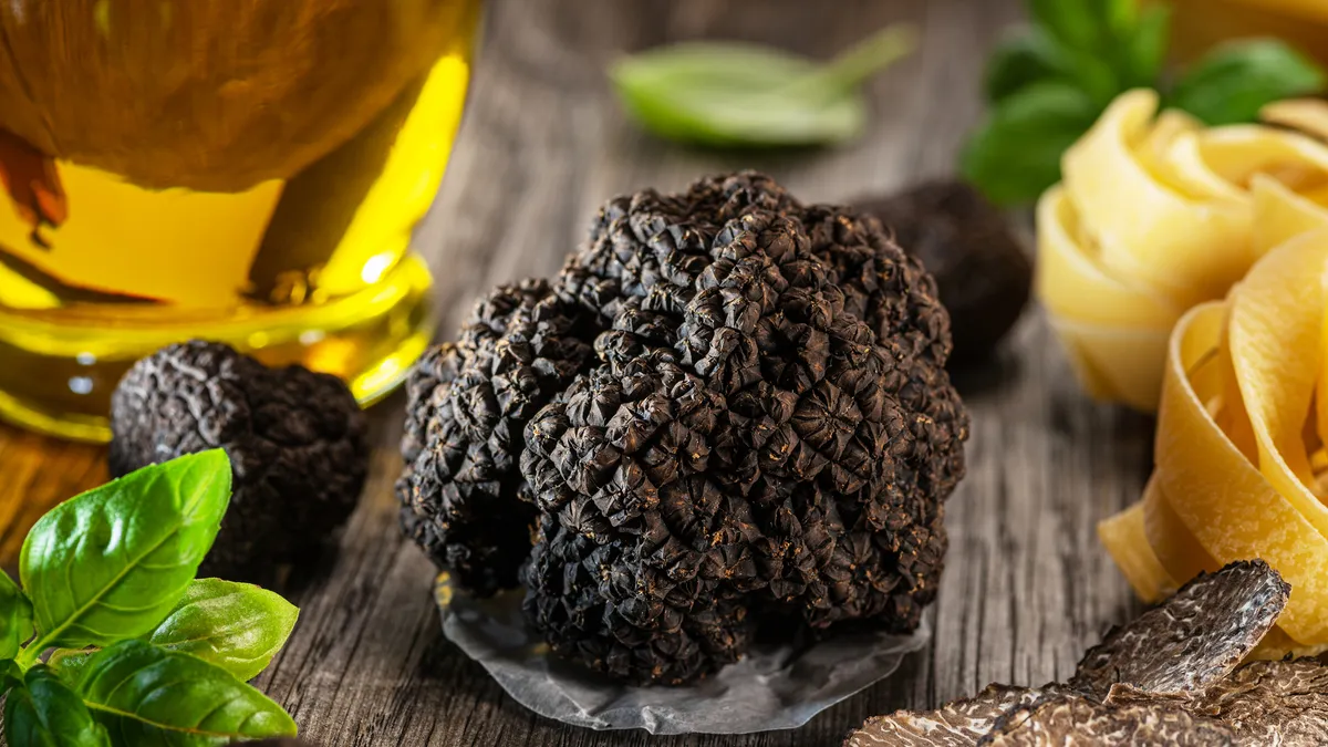 A truffle sits in the center of a wooden table surrounded by pasta, truffle shavings, basil and oil.