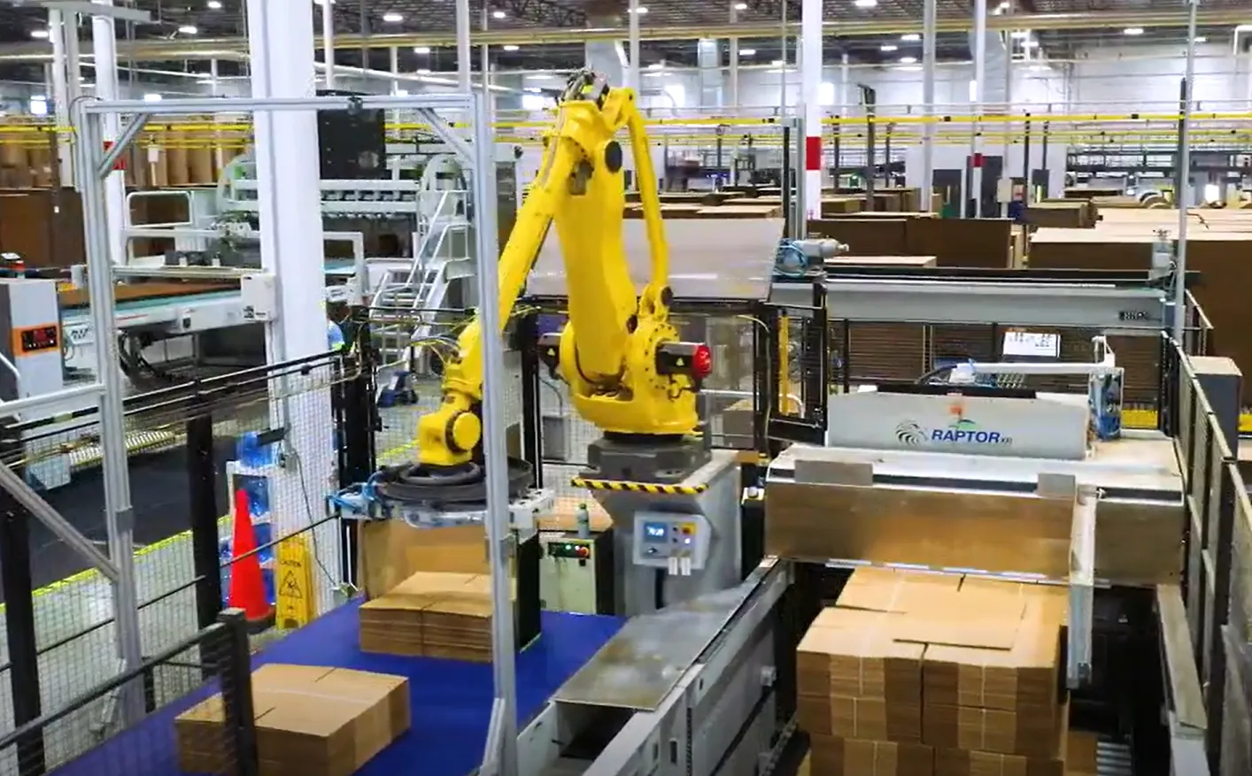 A yellow robotic arm stacks bundles of corrugated boxes in an industrial facility.