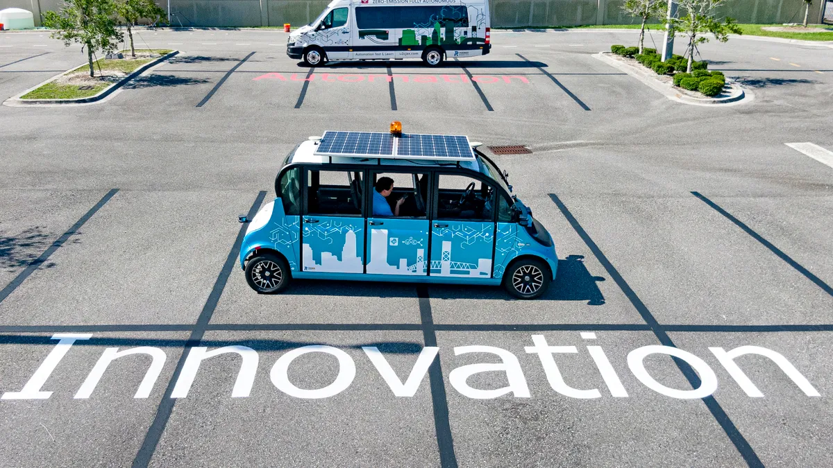 Looking down at an angle, showing a small four-wheel self-driving vehicle with a solar panel on the roof and a passenger in the second-row seat looking at their cell phone.