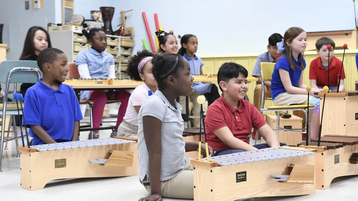 A group of students are sitting on the floor and in seats in a classroom. Music instruments are in front of each of them.