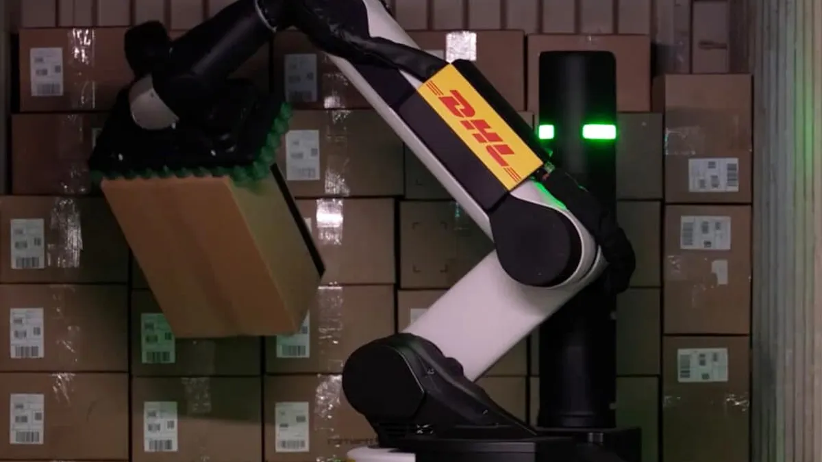 A robot arm with the DHL logo on it moves a brown cardboard box in front of a stack of other boxes.
