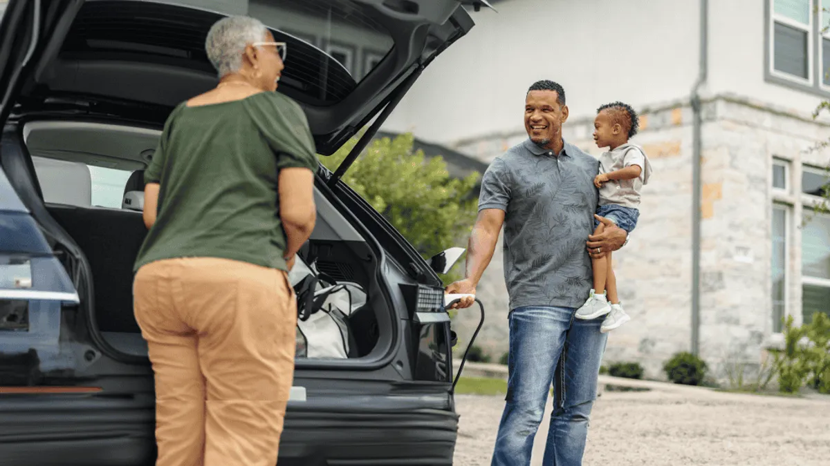 A family packs their car for a trip, with a man holding a child and a woman loading the trunk.