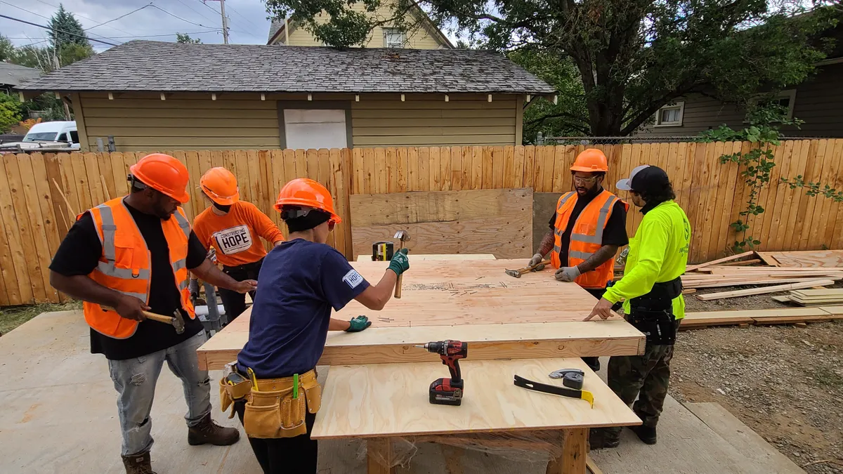 A group of five workers in construction gear hammer nails into a project on a workstation.