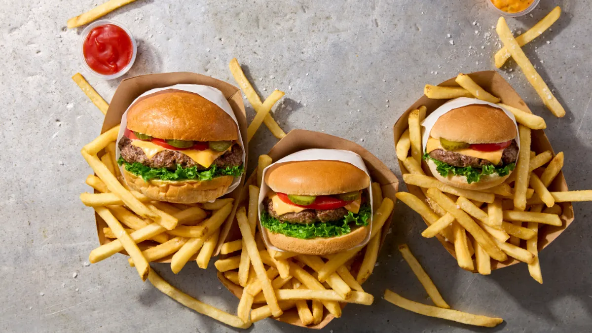 Overhead view of three cheeseburgers in paper trays, surrounded by golden French fries with ketchup and dipping sauce on a gray stone surface.