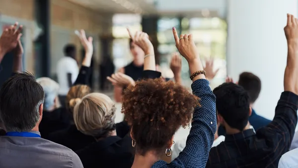 From behind: Several people in a group are raising their hands; in focus, a curly-haired person with hoops and brown skin