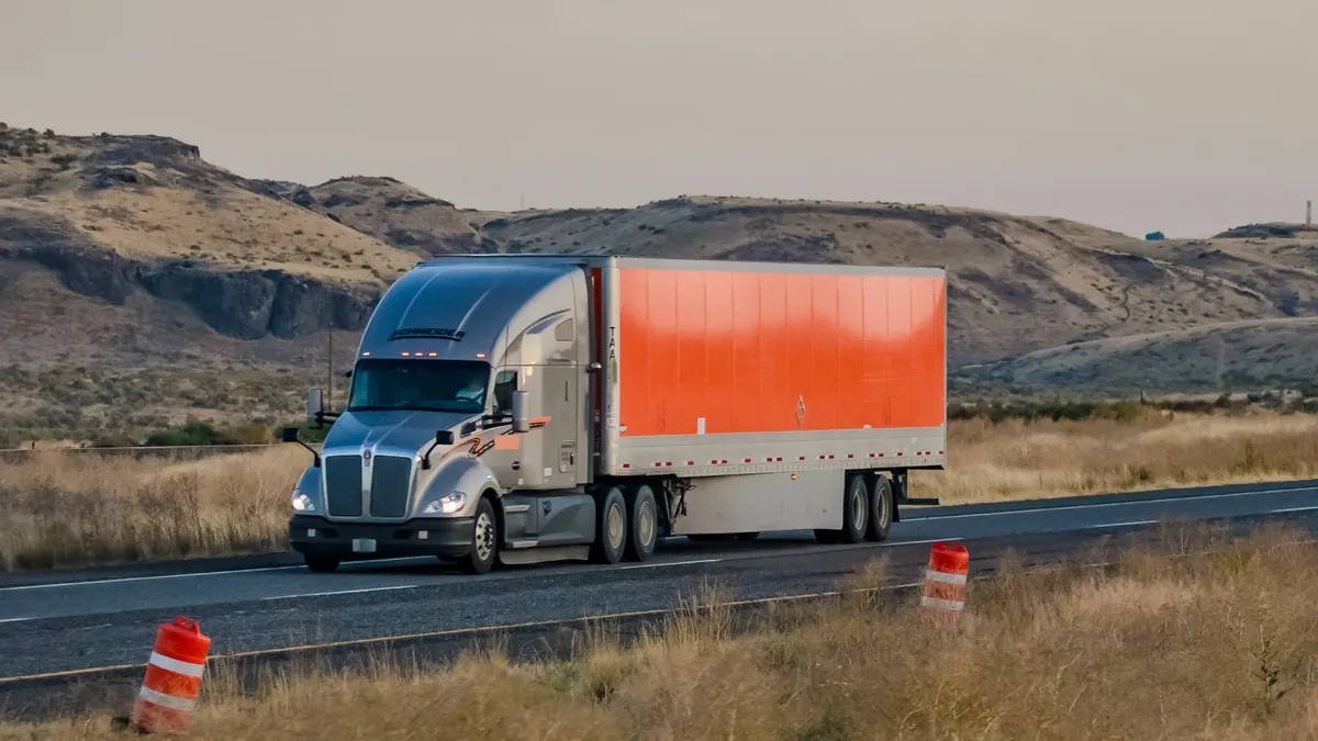 A Schneider National tractor-trailer traveling on US-395 in Washington state with large construction barrels on the side of the roadway.