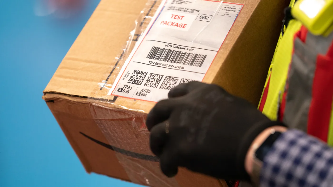 A person wearing black gloves holds a cardboard e-commerce box with QR codes and a barcode printed on the address label.