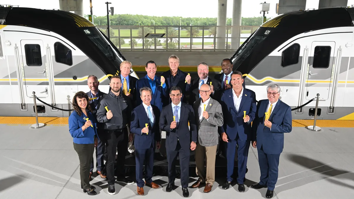 A group of 12 men and 1 woman in suits holding yellow rail spikes stand on a platform between two trains.