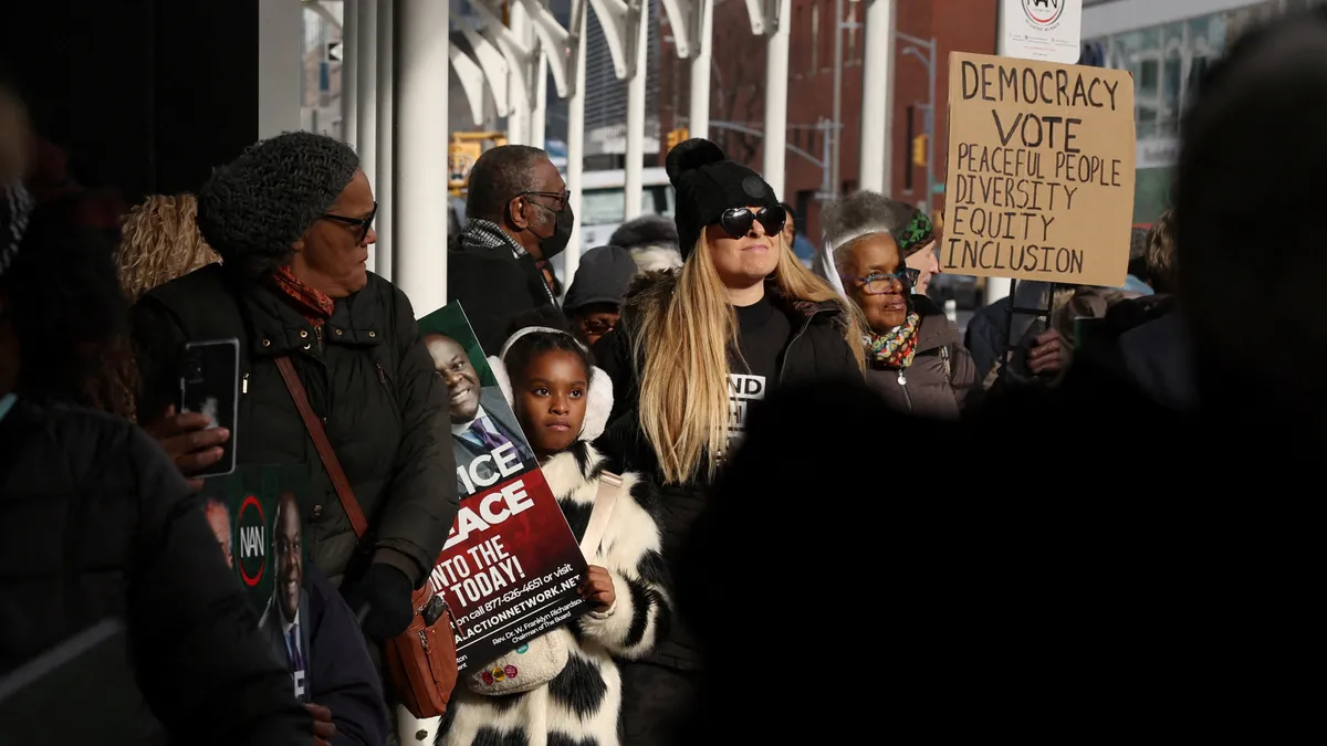 A crowd of people, representing a mix of genders and ethnicities, hold signs advocating for DEI.