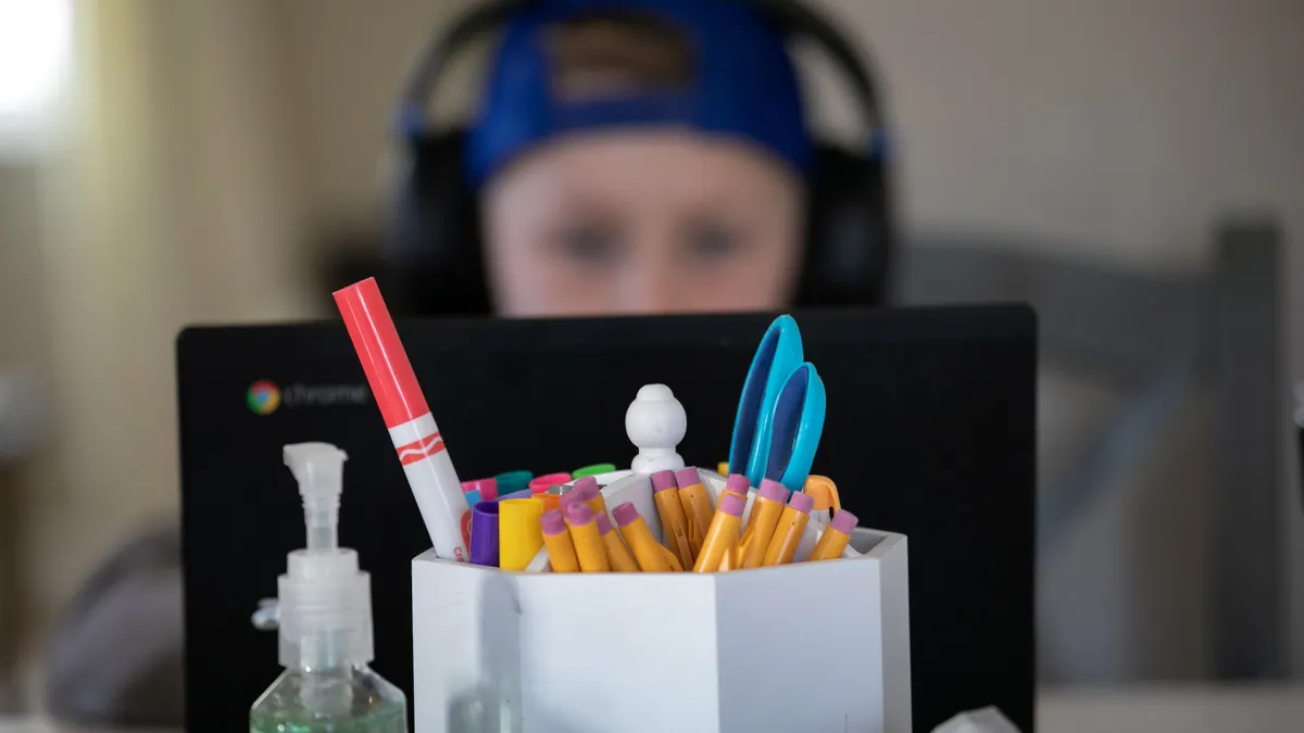 A young student wearing headphones works on a laptop behind a focused image of school supplies and a bottle of hand sanitizer.