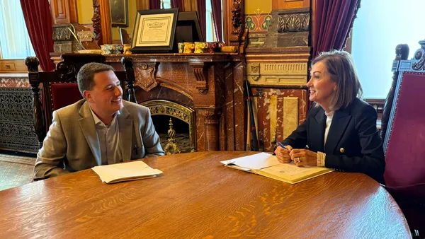 Two people in professional business attire sit at a wooden desk and discuss the contents of the legal documents in front of them.