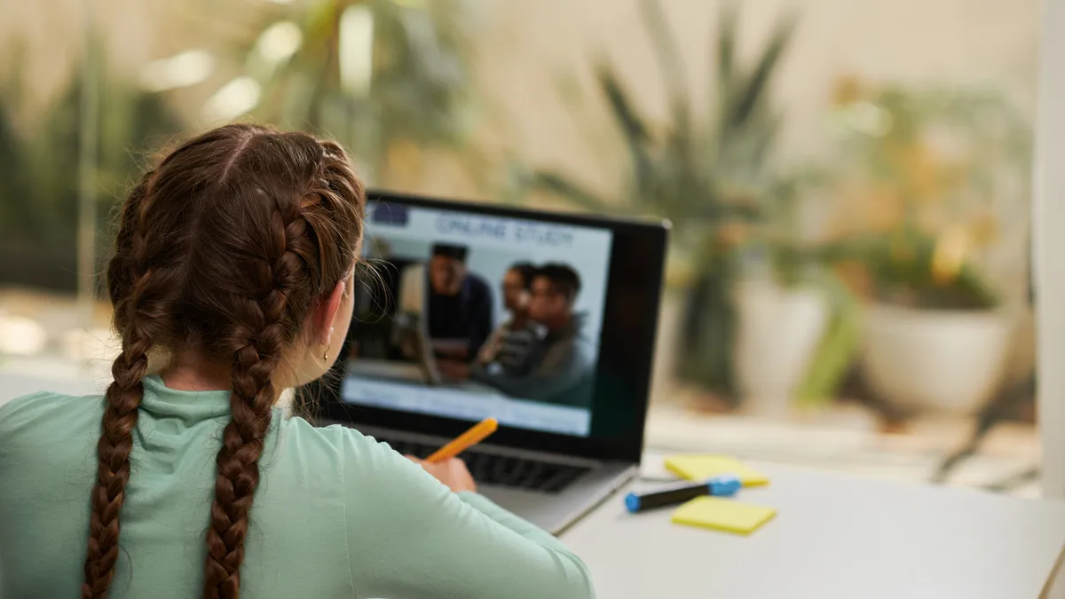 A young student watches a remote lesson on their computer while taking notes at their desk.